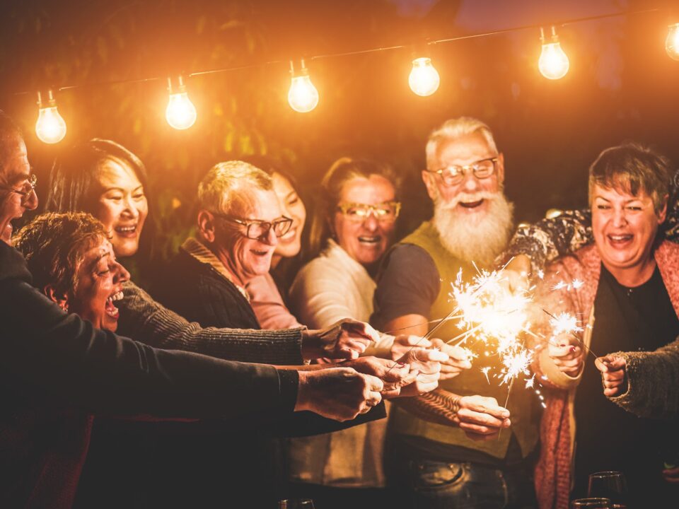 Many friends are gathered under warm outdoor lights with smiles on their faces, holding lit sparklers in celebration.