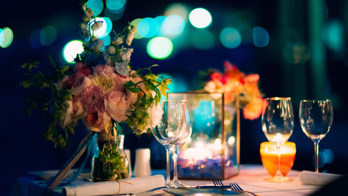 This is a darkened image with a tablescape featuring candles, various clear glasses for wine and other drinks, a white table cloth and several floral bouquets.